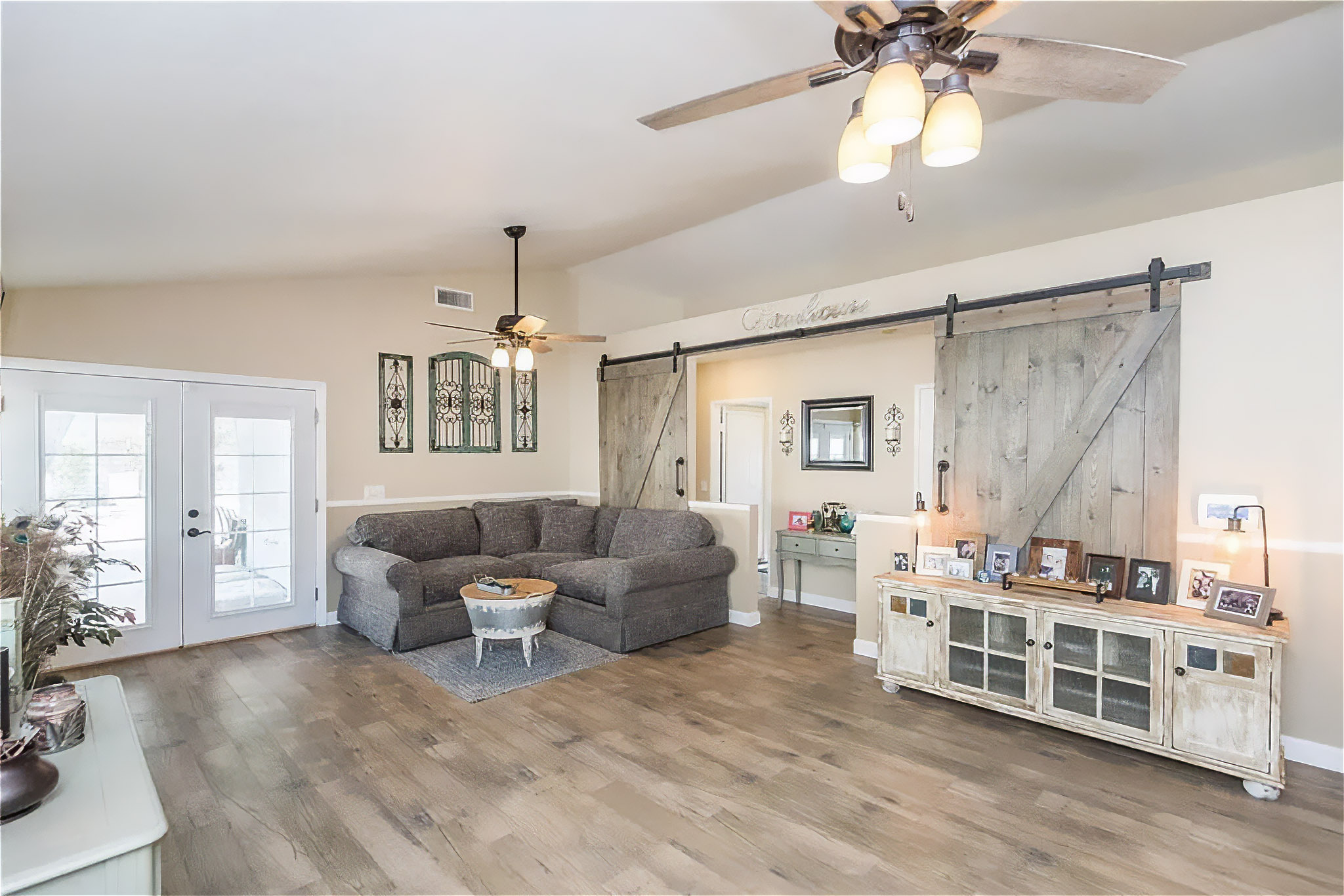 A bright, open living room with wood-look flooring and neutral beige walls. A gray sectional sofa sits in the corner near glass French doors that open to an outdoor space. Two ceiling fans with light fixtures hang from the ceiling. On the right, rustic sliding barn doors frame a wide opening to another room, with a distressed wood media console beneath family photos and decor. The space has a clean, farmhouse-inspired style with warm, natural light.
